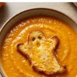 A bowl of orange butternut squash soup topped with a grilled cheese sandwich cut into a ghost shape.